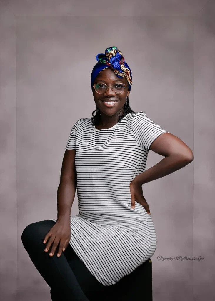 Emmy Mawumenyo seated in a striped dress, glasses and a blue head wrap, one hand on her hip.